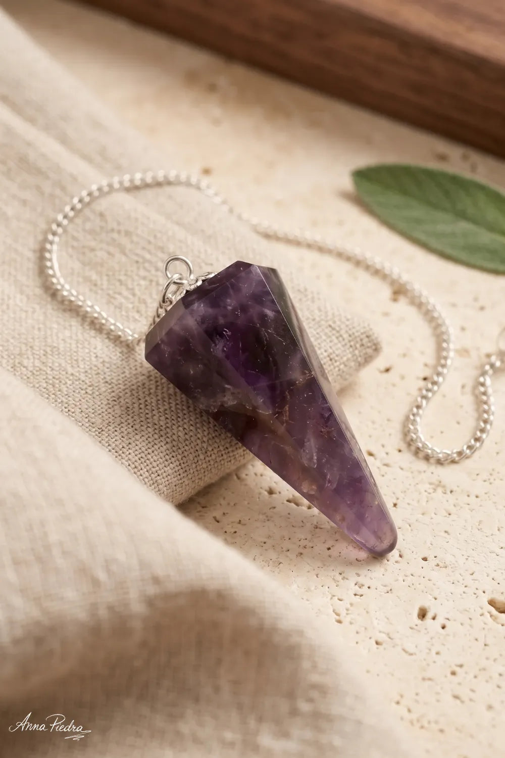 Primer plano de un Péndulo de Amatista pulida con tonalidades violetas y cadena plateada sobre tela de lino beige.