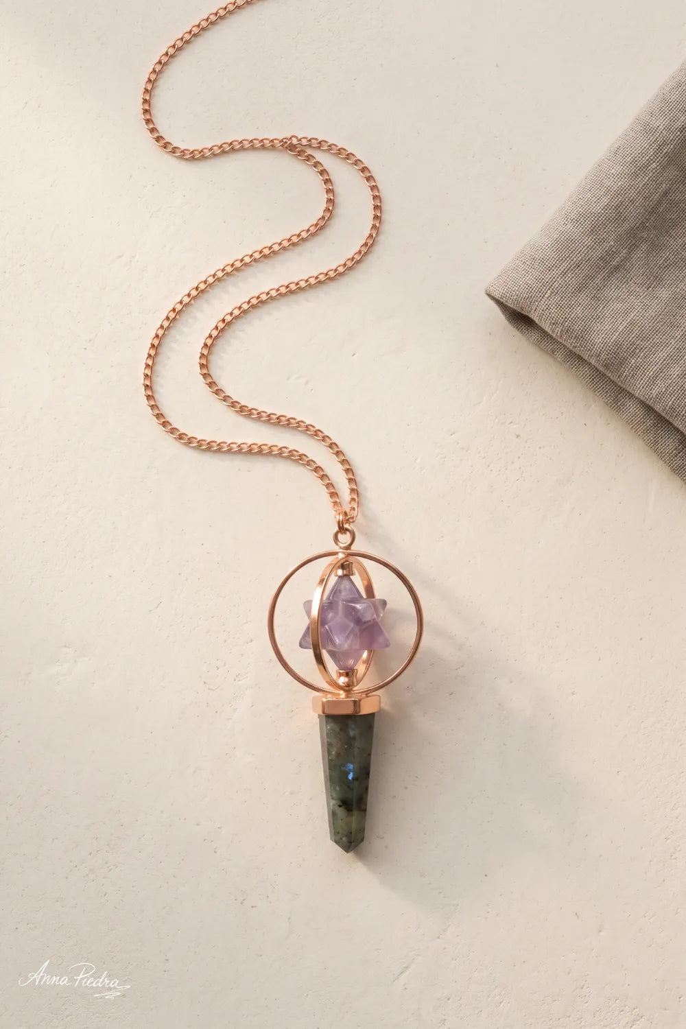 Péndulo Merkaba de amatista y labradorita con cadena color cobre extendida sobre una superficie beige.