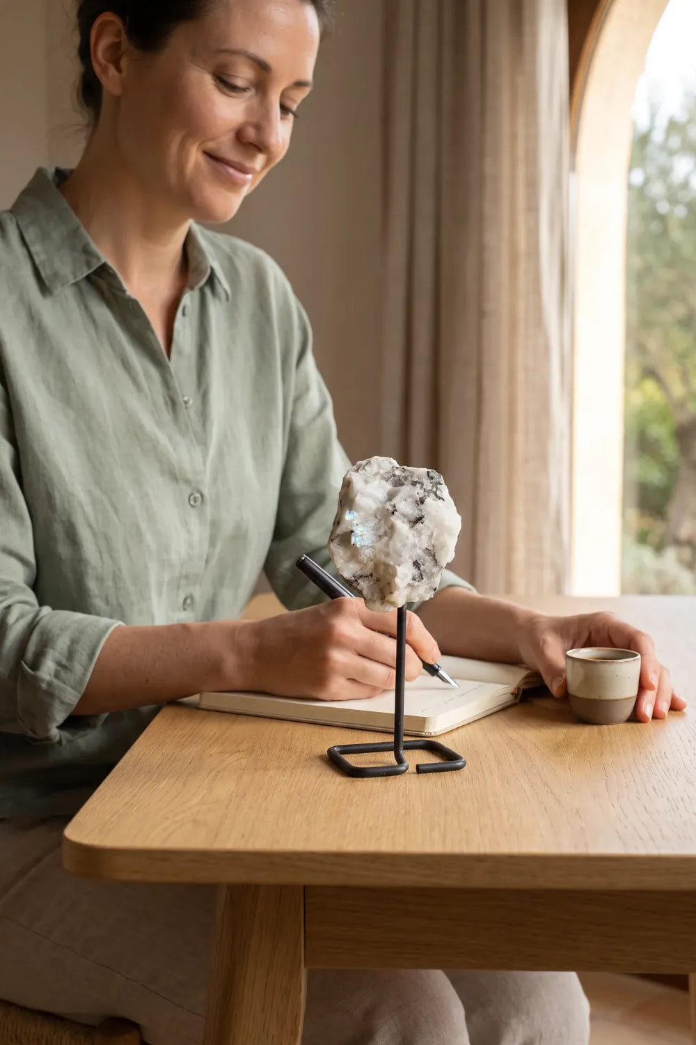 Mujer escribiendo en una libreta junto a una piedra luna en bruto con soporte de metal sobre una mesa de madera