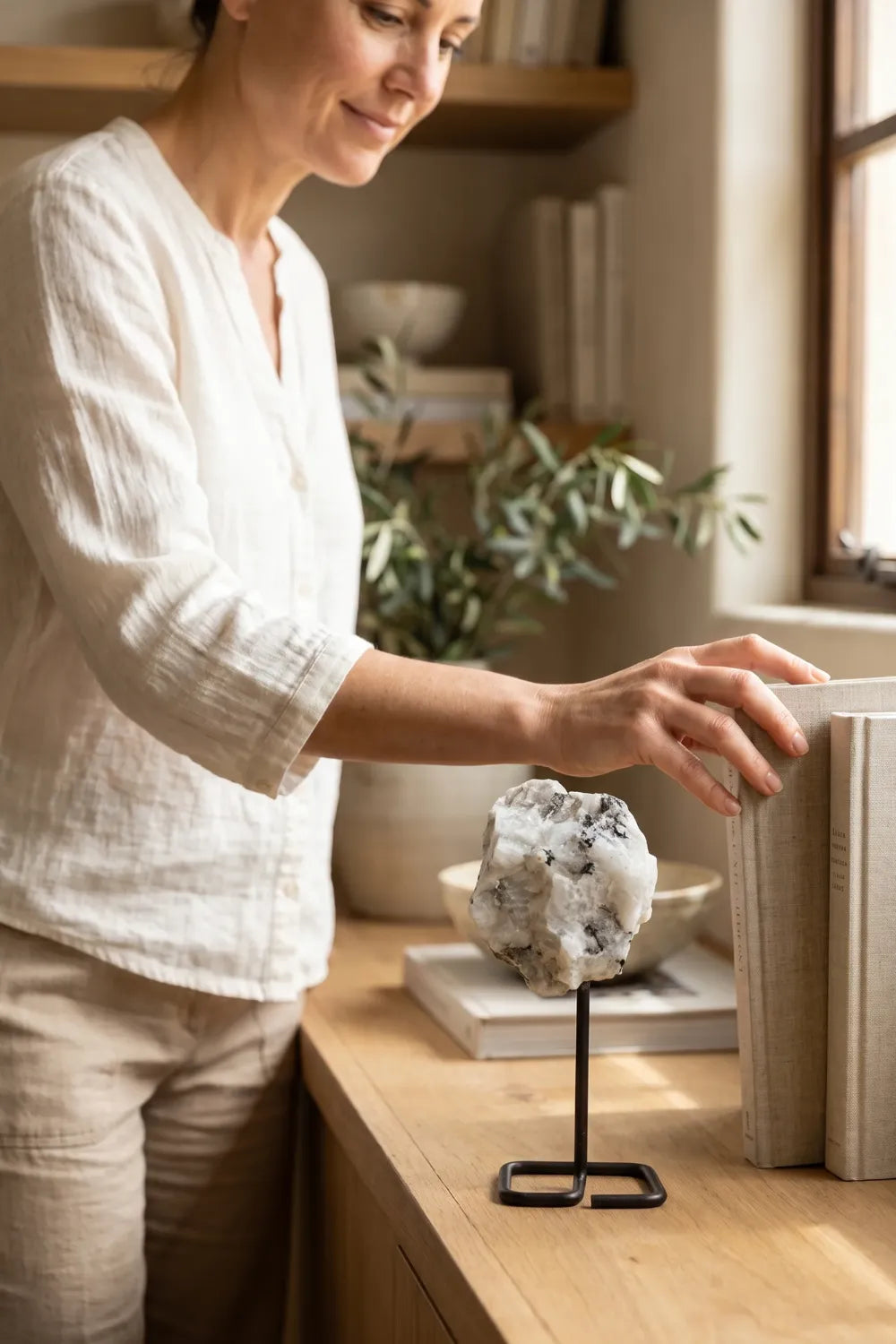 Mujer colocando libros junto a una piedra luna en bruto con soporte de metal sobre un mueble de madera