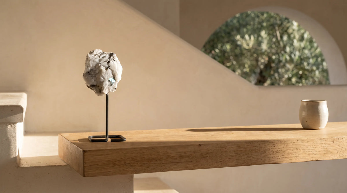 Piedra luna en bruto con soporte de metal sobre una repisa de madera, con fondo mediterráneo y pequeña vasija cerámica