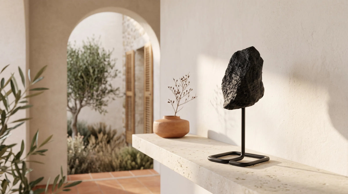 Piedra de turmalina negra natural en bruto sobre soporte metálico, expuesta en una estantería de piedra clara con decoración mediterránea.