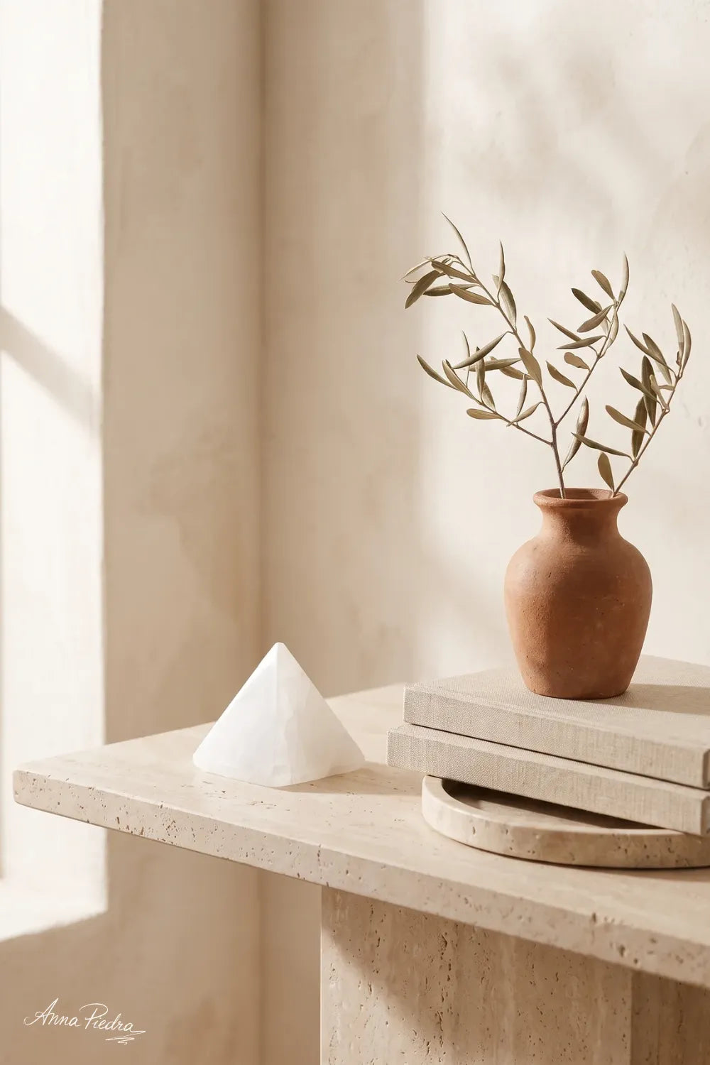 Pirámide de selenita natural sobre mesa de travertino junto a libros y jarrón con rama de olivo - Anna Piedra