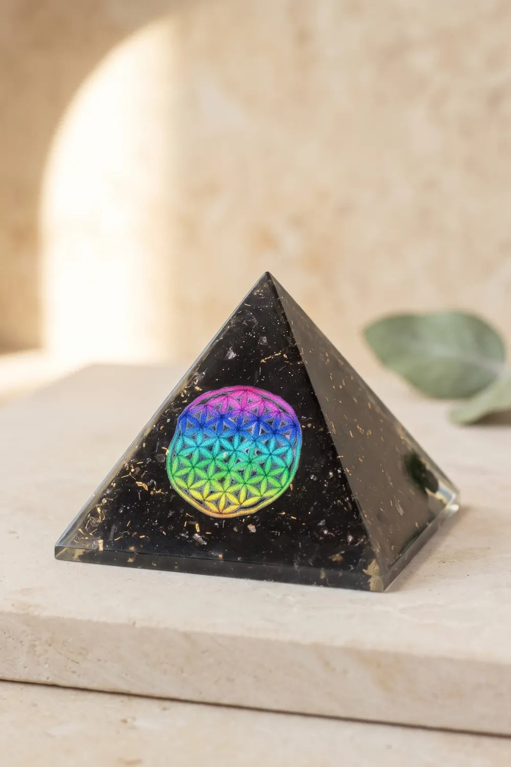 Primer plano de una pirámide de orgonita negra con partículas doradas y símbolo de la Flor de la Vida en colores arcoíris.