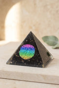 Primer plano de una pirámide de orgonita negra con partículas doradas y símbolo de la Flor de la Vida en colores arcoíris.