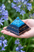 Pirámide de orgonita grande de turquesa y turmalina negra sostenida sobre la mano en un jardín con flores azules. anna piedra