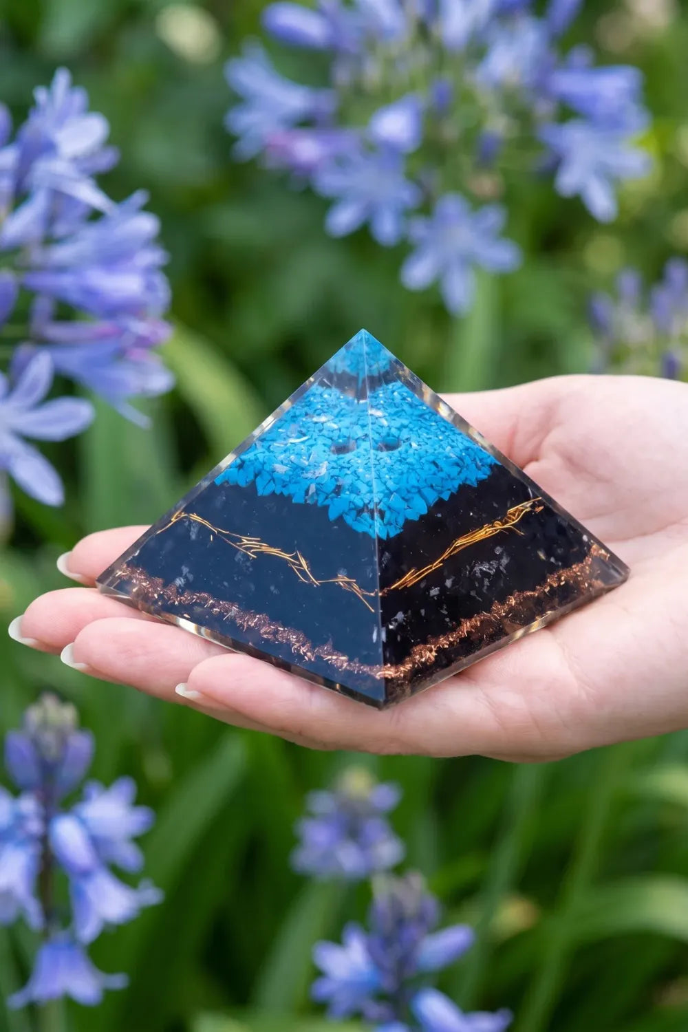 Pirámide de orgonita grande de turquesa y turmalina negra sostenida sobre la mano en un jardín con flores azules. anna piedra