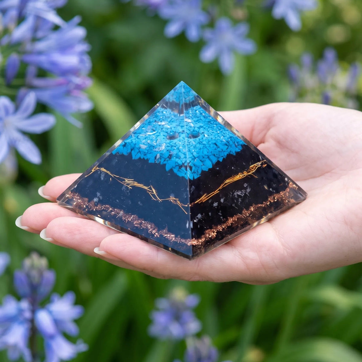 Pirámide de orgonita grande con turquesa y turmalina negra sostenida en la mano, con flores azules al fondo.