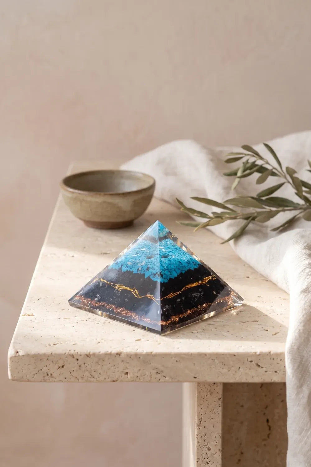 Pirámide de orgonita grande de turquesa y turmalina negra sobre una mesa de piedra clara con rama de olivo. anna piedra