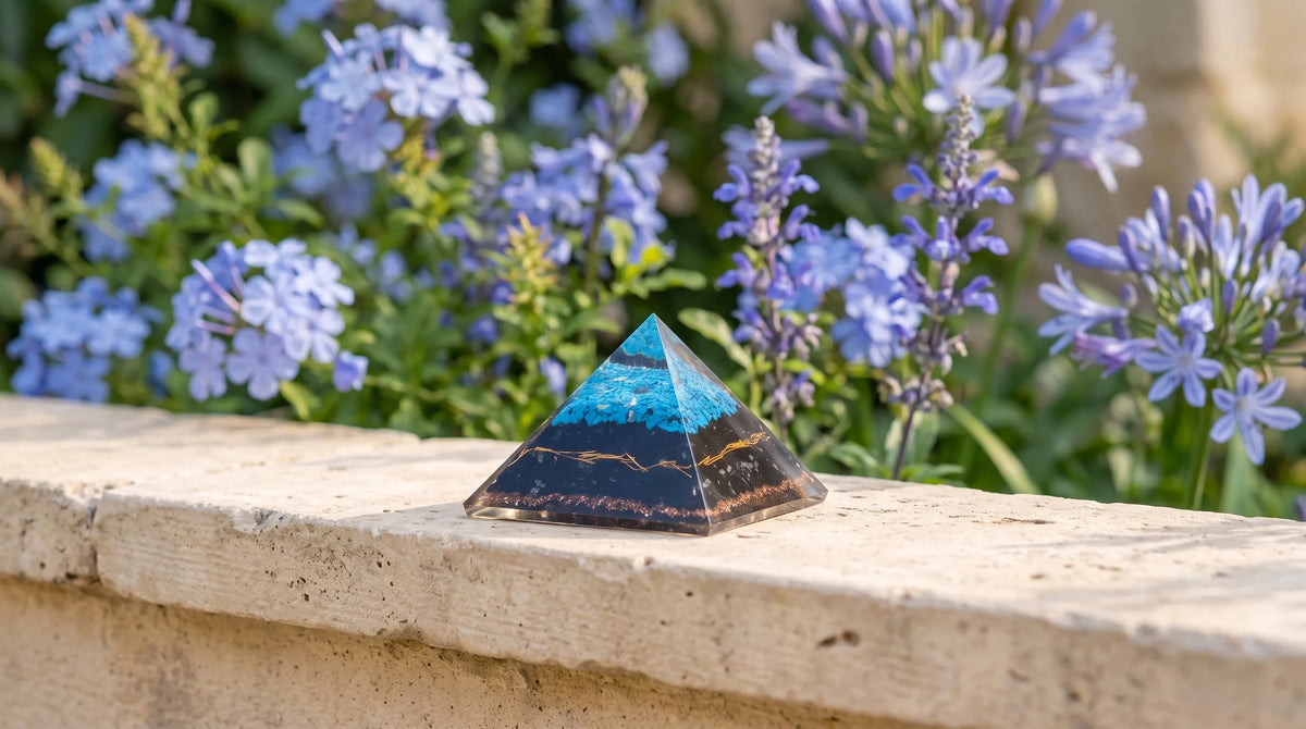 Pirámide de orgonita grande de turquesa y turmalina negra sobre un murete de piedra, con flores azules al fondo anna piedra