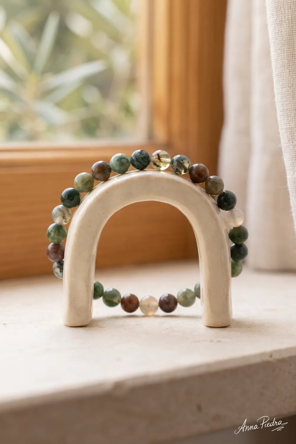 Primer plano de pulsera de ágata musgosa con cuentas pulidas verdes, marrones y translúcidas, sobre un soporte de cerámica beige junto a una ventana – Anna Piedra