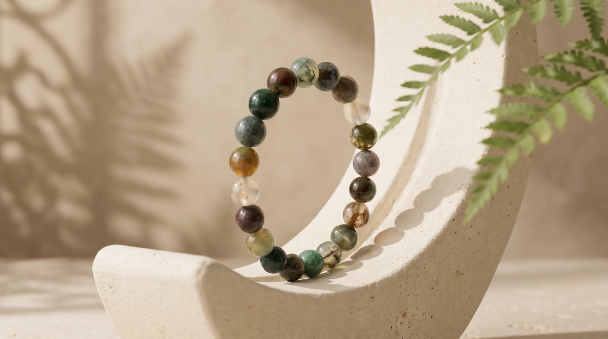 Pulsera de ágata musgosa natural con cuentas redondas verdes, marrones y translúcidas, colocada sobre un soporte curvo de piedra beige – Anna Piedra