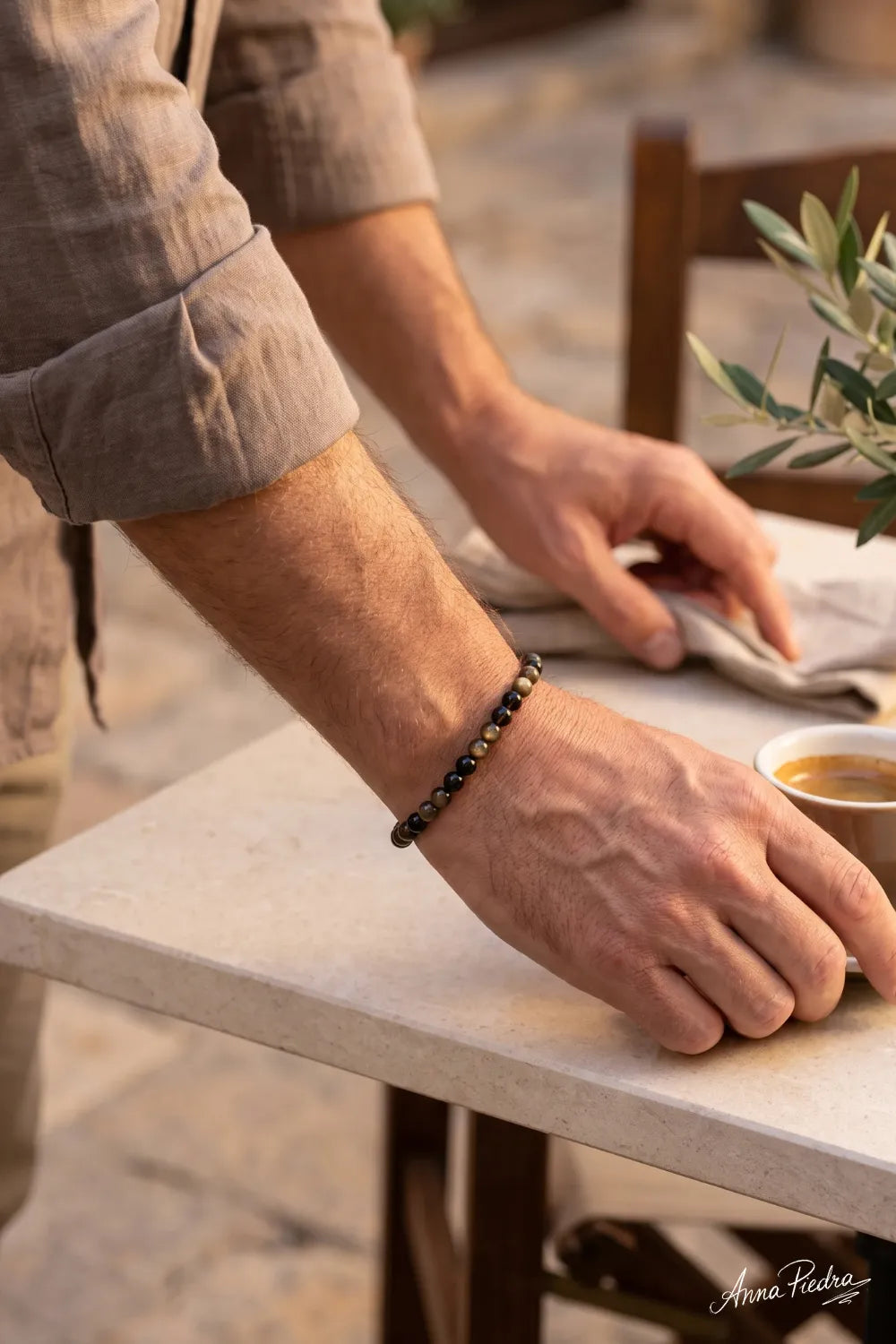 Pulsera de obsidiana dorada para hombre con cuentas de 6 mm y reflejos dorados, detalle en la muñeca junto a una mesa de café – Anna Piedra