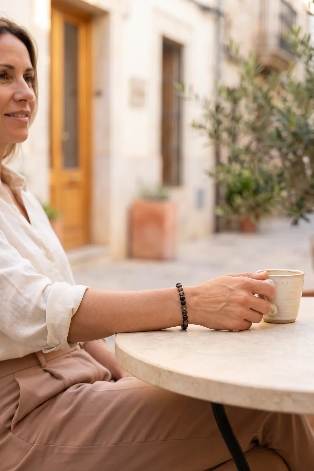 Mujer con pulsera de obsidiana dorada en la muñeca mientras sostiene una taza de café en una terraza mediterránea – Anna Piedra