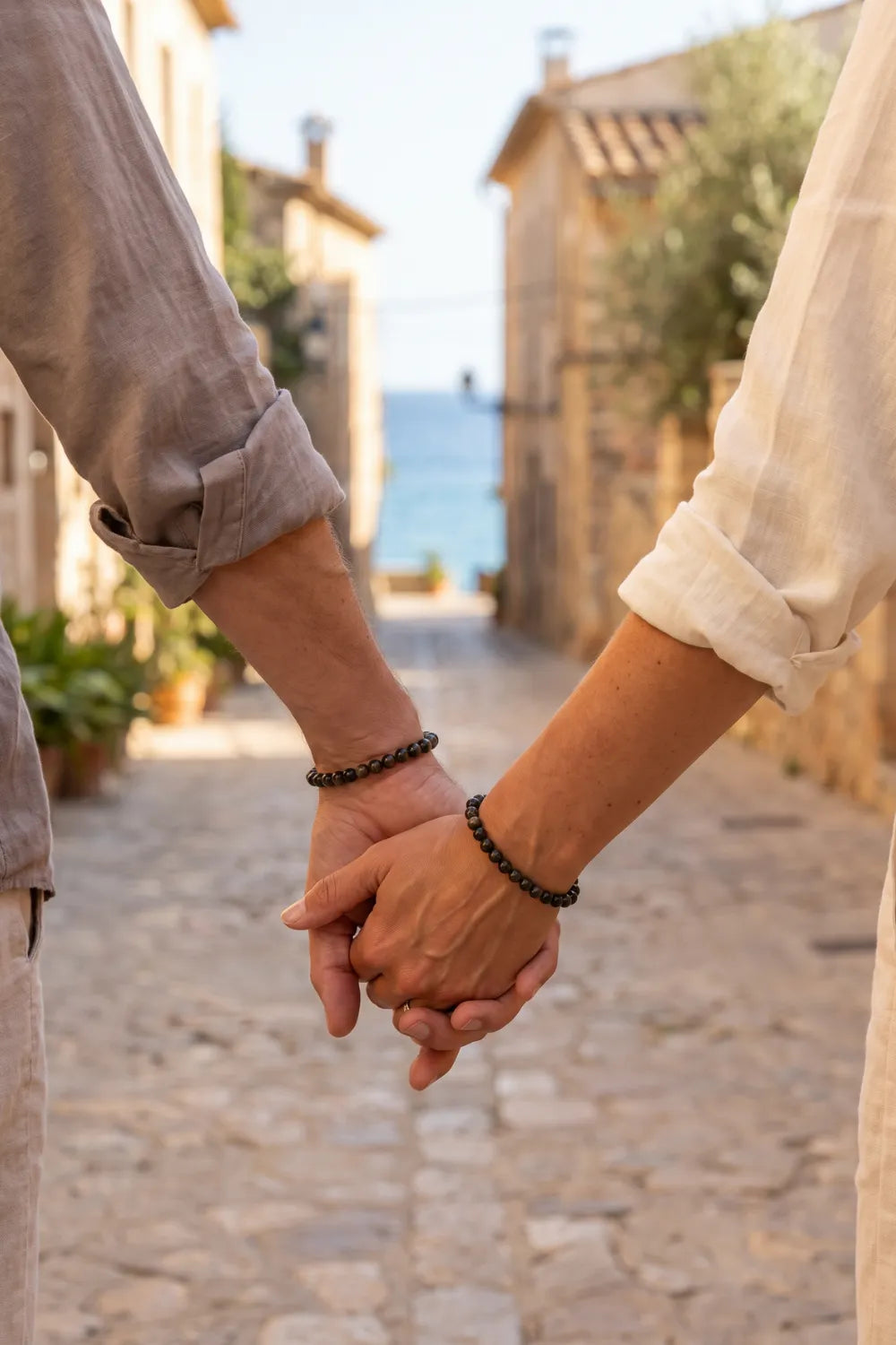 Pareja cogida de la mano con una pulsera de obsidiana dorada de cuentas negras y doradas en cada muñeca, en una calle mediterránea – Anna Piedra