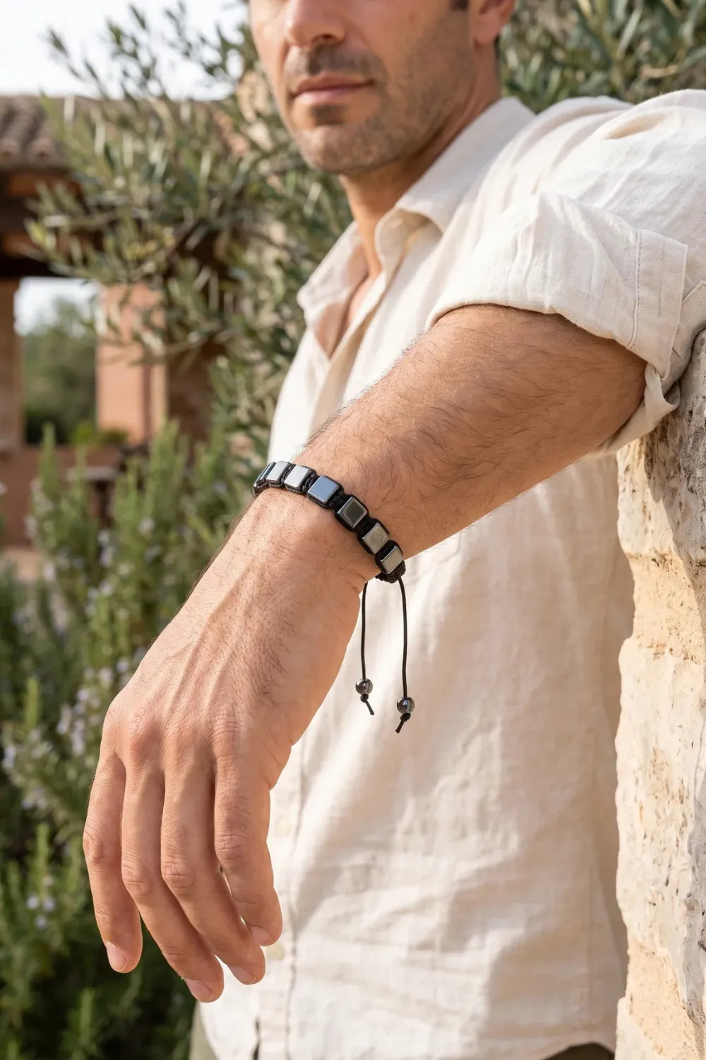 Primer plano de una pulsera de hematita shamballa ajustable en la muñeca de un hombre junto a un muro de piedra. anna piedra