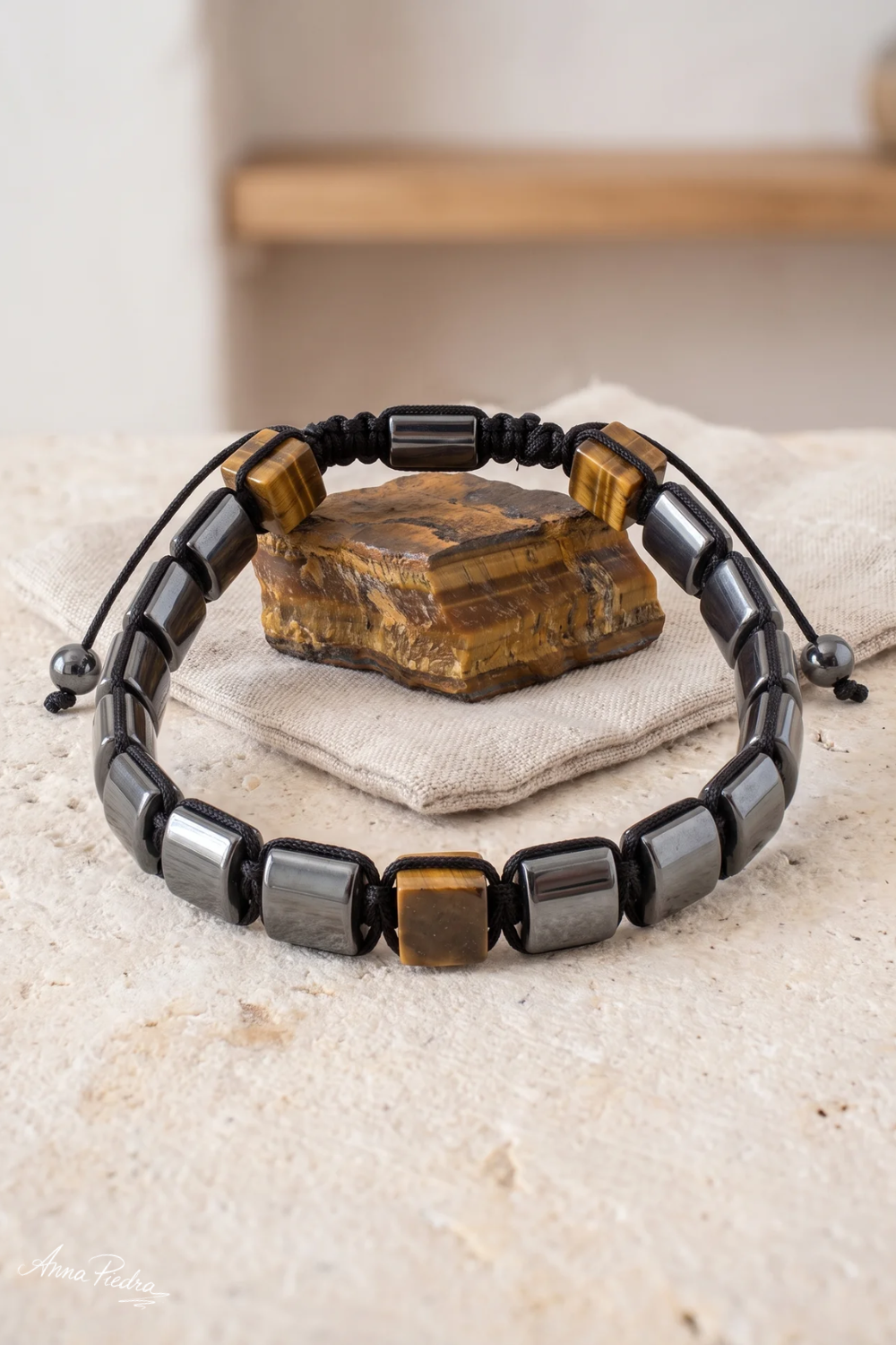 Pulsera de hematita y ojo de tigre con cuentas cuadradas y cordón negro junto a una piedra de ojo de tigre en bruto. anna piedra