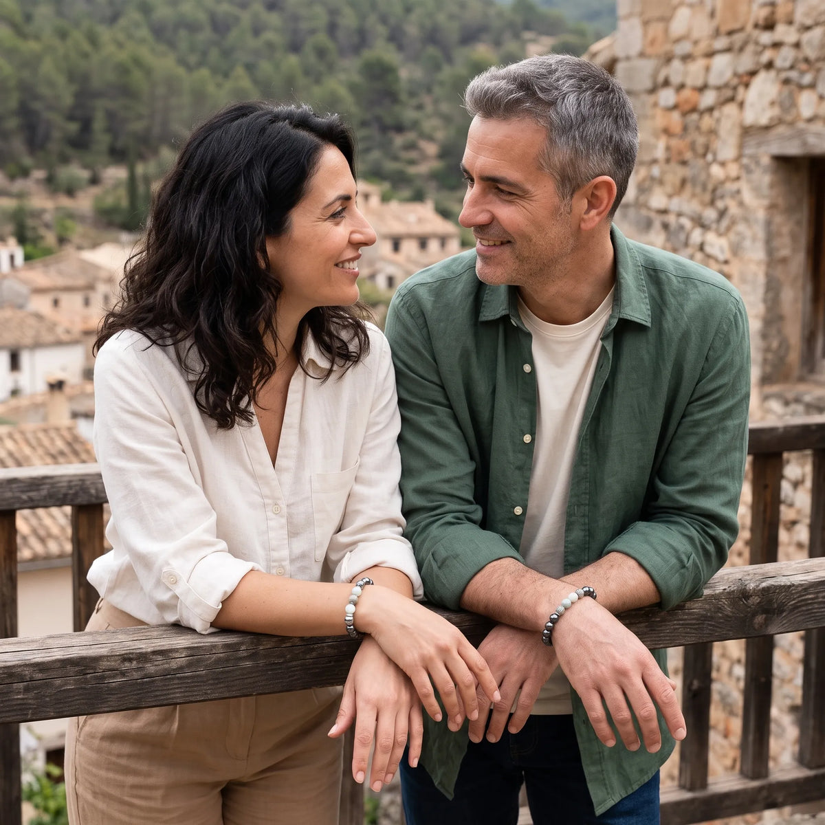 Pareja sonriendo en un balcón de pueblo mediterráneo luciendo pulseras de jade de montaña y hematita.