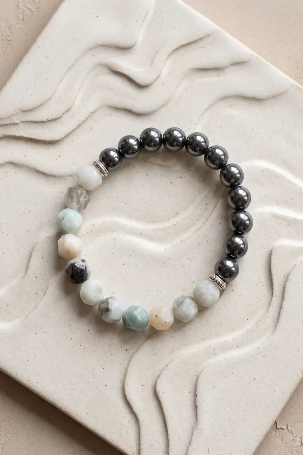 Pulsera de jade de montaña y hematita sobre una placa de cerámica clara con relieve ondulado.
