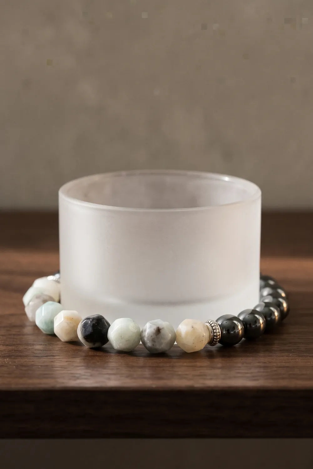 Pulsera de jade de montaña y hematita rodeando un portavelas blanco sobre una superficie de madera oscura.