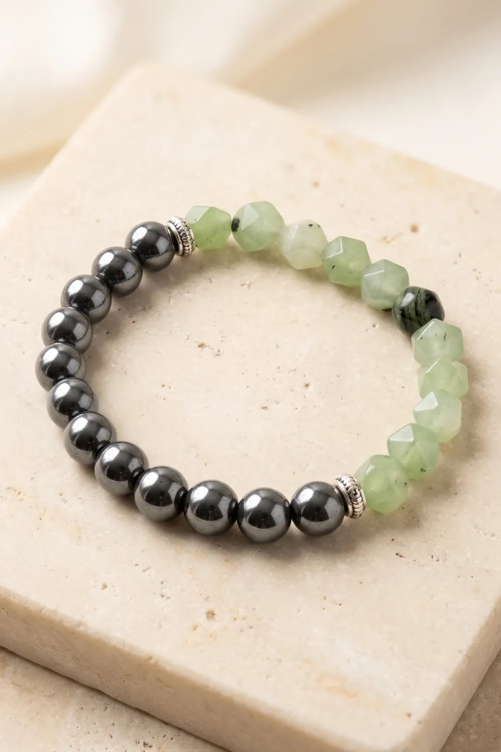 Primer plano de una pulsera de jade verde con cuentas facetadas, hematita pulida y separadores plateados sobre piedra natural.