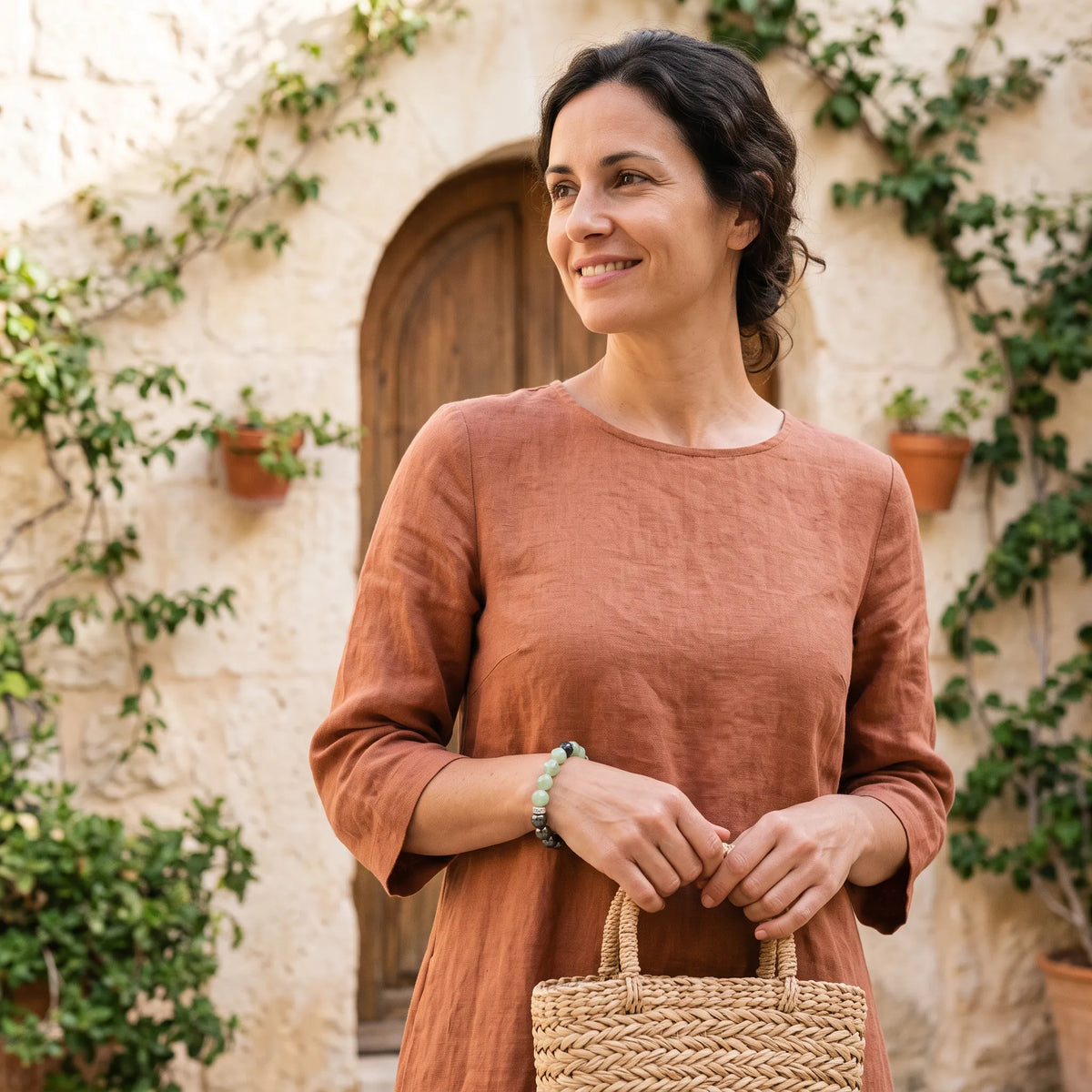Mujer con vestido terracota y cesta de mimbre luciendo una pulsera de jade verde y hematita en un patio mediterráneo.