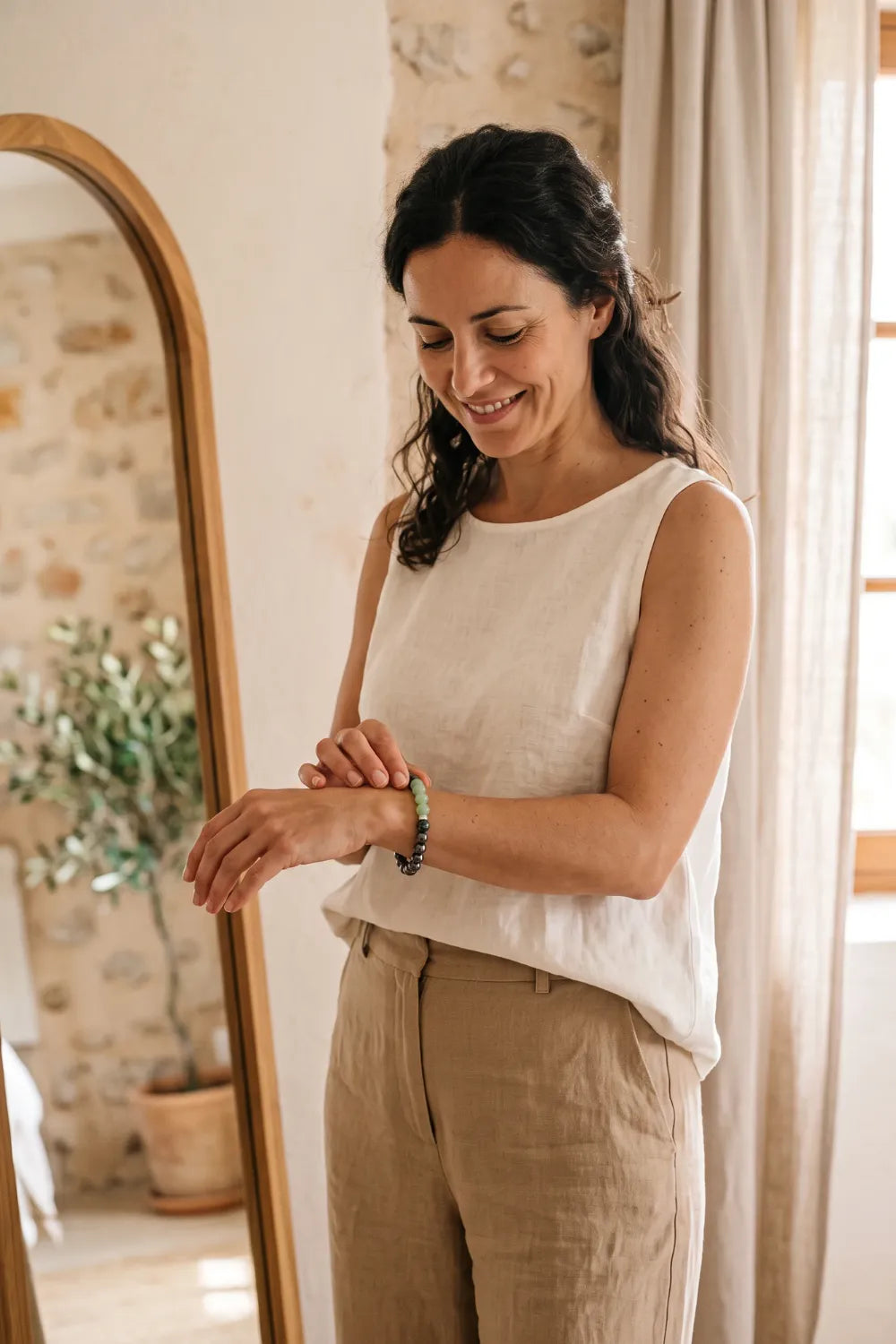 Mujer con ropa de lino blanco ajustándose una pulsera de jade verde y hematita junto a un espejo de madera.