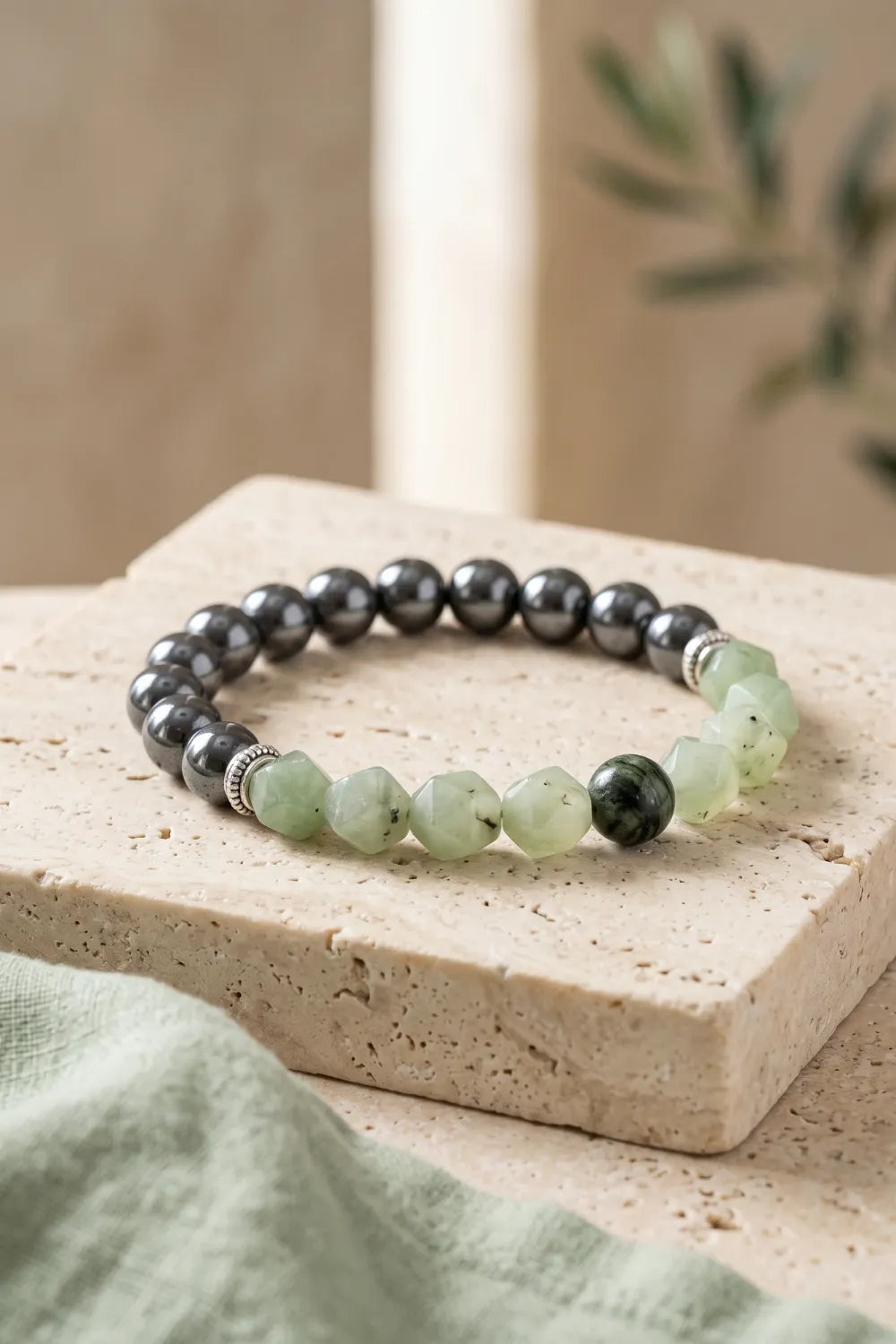 Pulsera de piedras naturales con cuentas de jade verde facetado y hematita gris metalizada sobre una base de piedra beige.