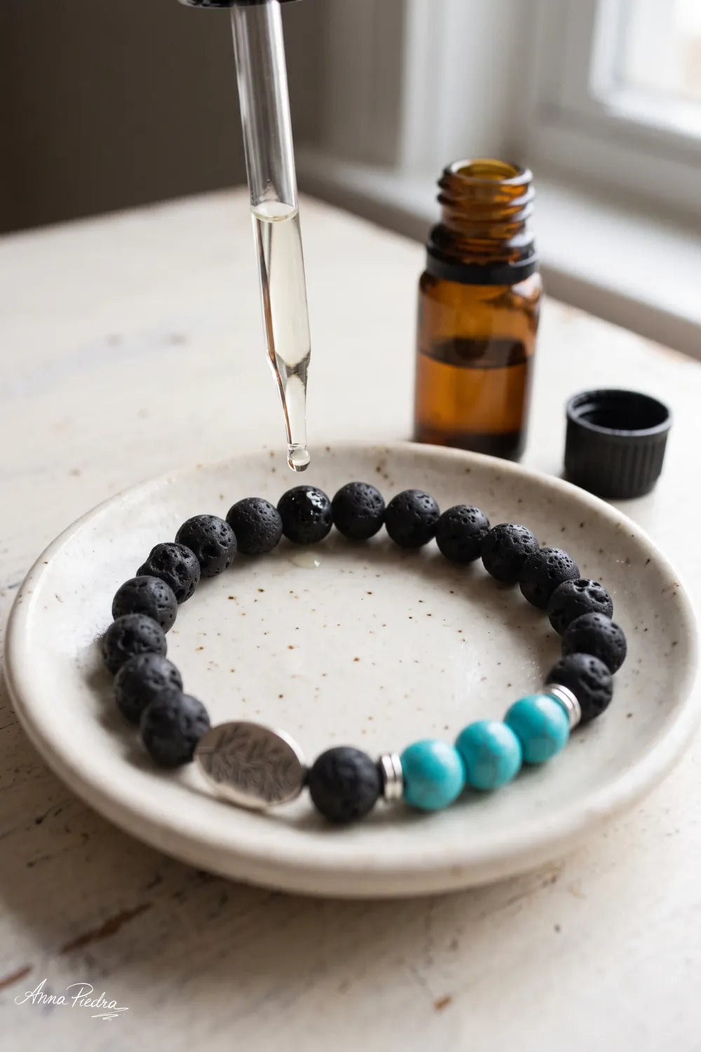 Pulsera de lava y turquesa en un plato de cerámica mientras una pipeta deja caer aceite esencial sobre las cuentas de lava.