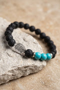 Pulsera de lava y turquesa con cuentas negras volcánicas, tres piedras turquesa y pieza ovalada plateada sobre una piedra clara.