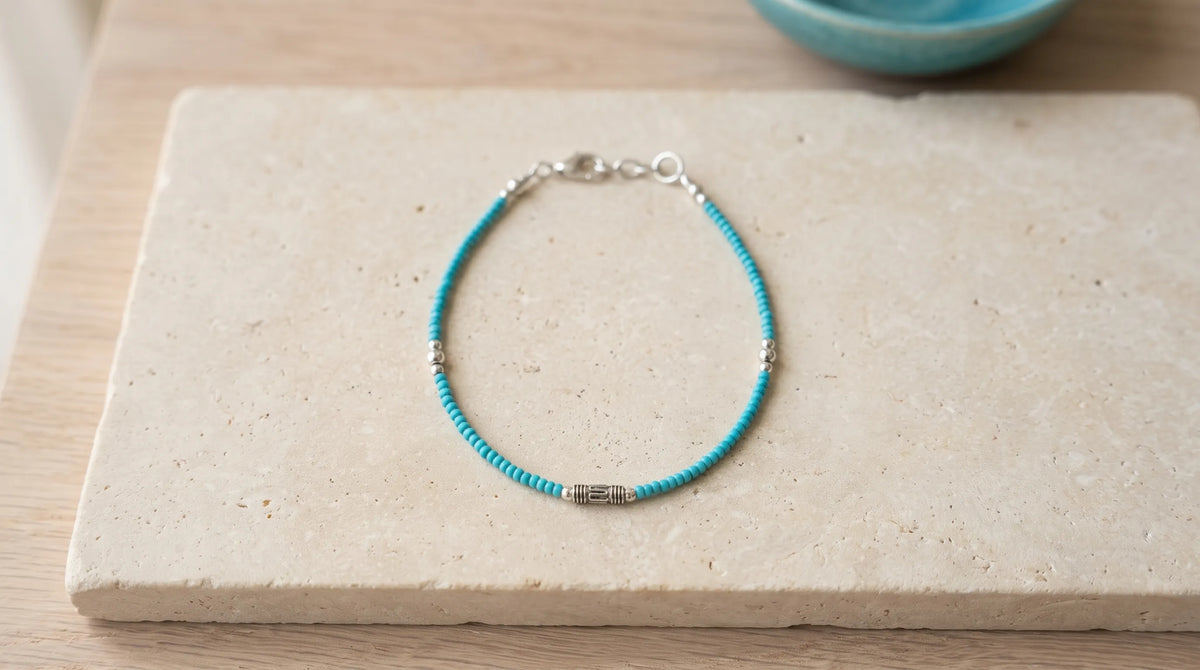 Pulsera de turquesa con pequeñas cuentas azules, detalles plateados y cierre ajustable sobre piedra beige.