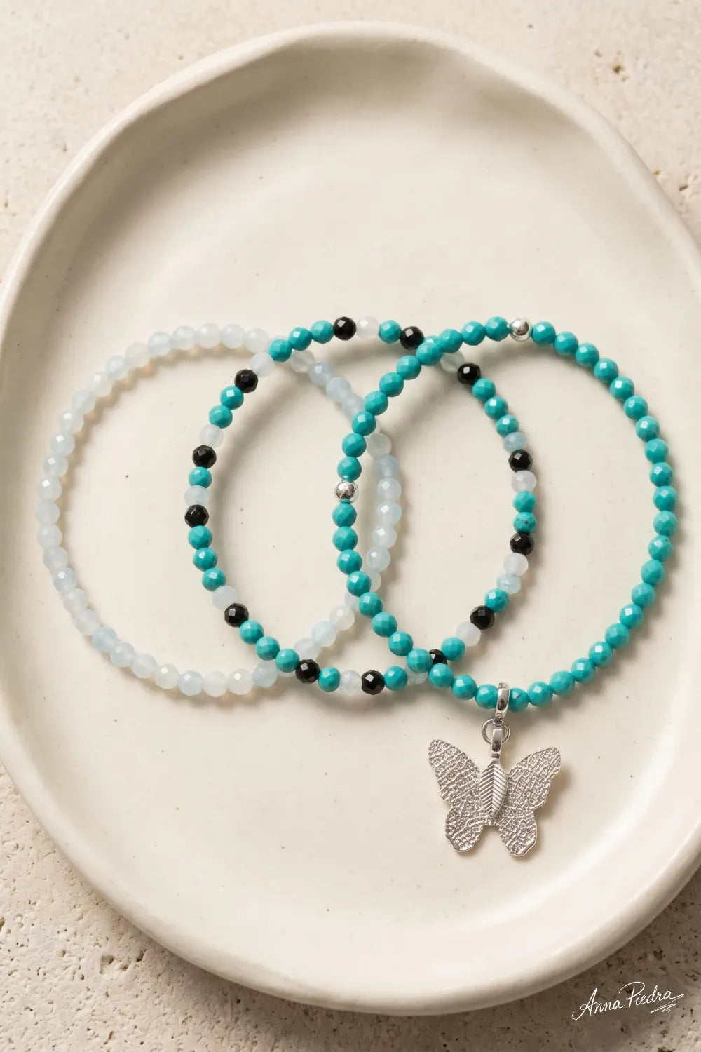 Conjunto de tres pulseras de cuentas turquesas, blancas y negras con colgante de mariposa plateada, colocado en una bandeja de cerámica blanca.