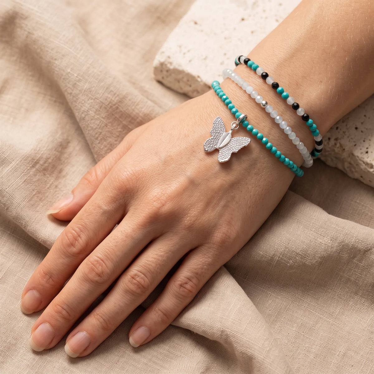 Primer plano de tres pulseras con cuentas turquesas, blancas y negras en la muñeca de una mujer, con un colgante de mariposa plateada.