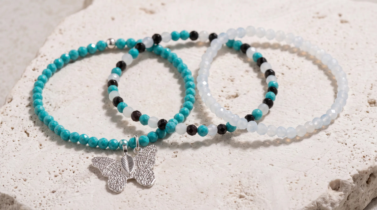 Set de tres pulseras con turquesas, cuentas blancas y negras y mariposa plateada, dispuestas sobre una superficie de travertino beige.
