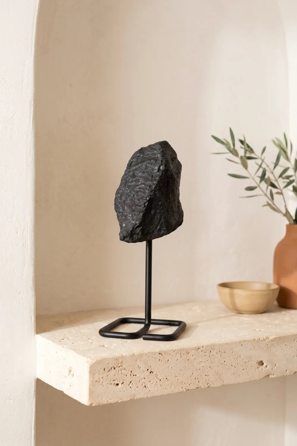 Piedra de turmalina negra en bruto con textura irregular y soporte negro, colocada en una repisa de piedra junto a una planta de olivo.